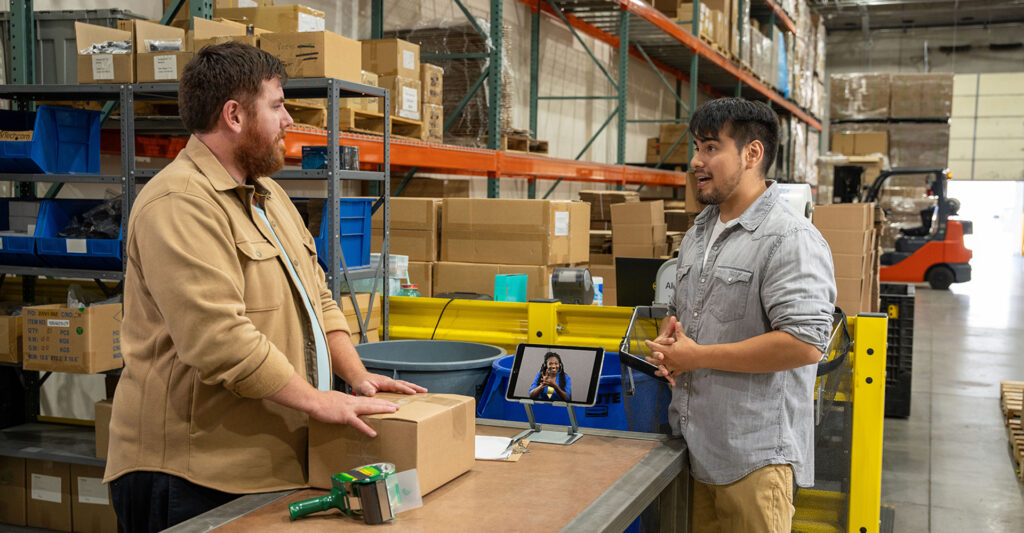 Coworkers in warehouse stand at packaging station. Mounted tablet between them shows live ASL interpreter.