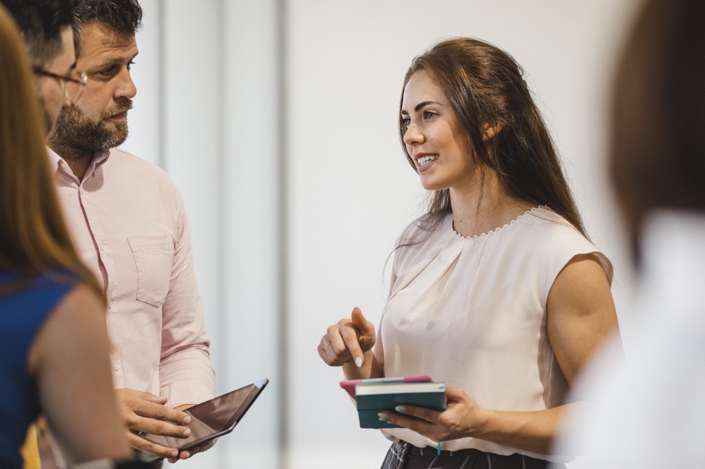 Businesswoman with long brown hair smiles while talking with two colleagues. She is holding a notebook in her hand.