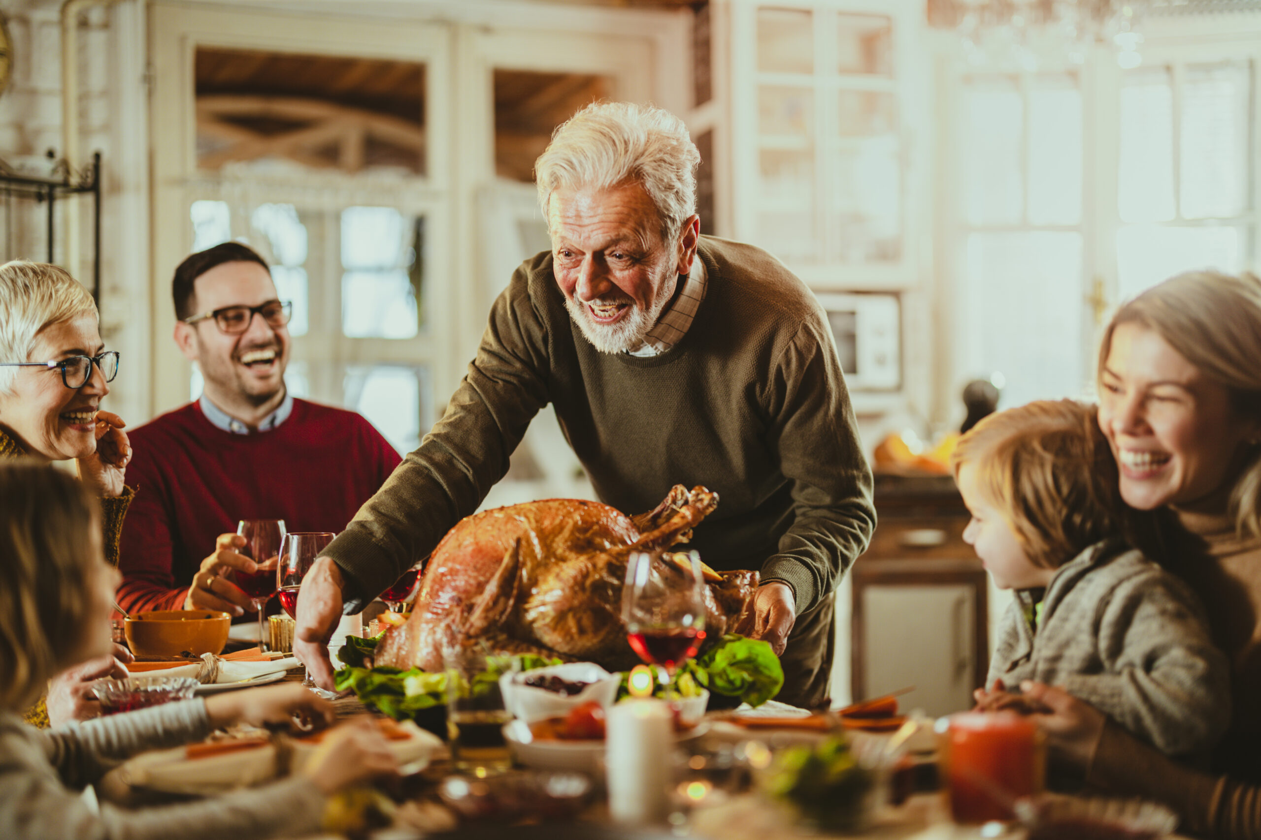 Happy senior man serving Thanksgiving turkey for his family at dining table.