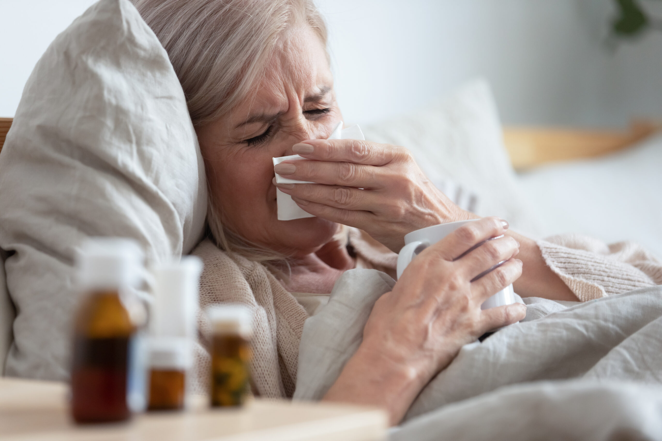 Ill middle aged woman sneezing blowing nose sit on bed Ill sick middle aged woman sneezing blowing running nose holding tissue sit on bed, upset old mature lady caught cold got flu influenza grippe symptoms drink hot tea taking medications at home alone